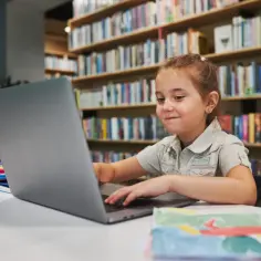 Dziewczynka uczy się na laptopie w bibliotece, szukając informacji, od której klasy jest geografia.