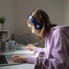 Dziewczynka w słuchawkach skupiona na nauce zdalnej, pisze w zeszycie przy laptopie.
