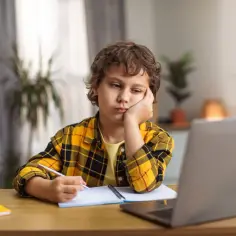 Chłopiec z czwartoklasistą znużony pisze wypracowanie. Na biurku laptop, zeszyt i przybory.