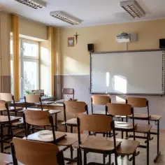 Pusta klasa z ławkami, tablicą interaktywną i krzyżem. Czy religia wlicza się do czerwonego paska? Słońce wpada przez okna.
