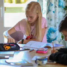 Dzieci odrabiają lekcje. Dziewczynka z blond włosami przegląda zeszyt, chłopiec pisze. Wygląda na to, że nie ma zadania domowego.