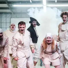 Aktorzy w białych strojach, z przerażonymi minami, tworzą scenę pełną grozy i komizmu w obrazie śmierci. Dym i teatralne światła podkreślają nastrój.
