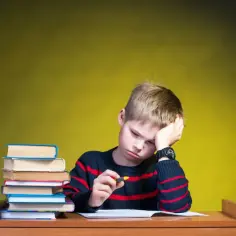 Zmęczony chłopiec z książkami przy biurku, z ręką na czole.