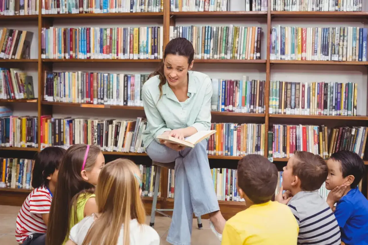 Nauczycielka czyta dzieciom w bibliotece, wprowadzając zmianę w podstawie programowej. Dzieci słuchają z uwagą, siedząc na podłodze.