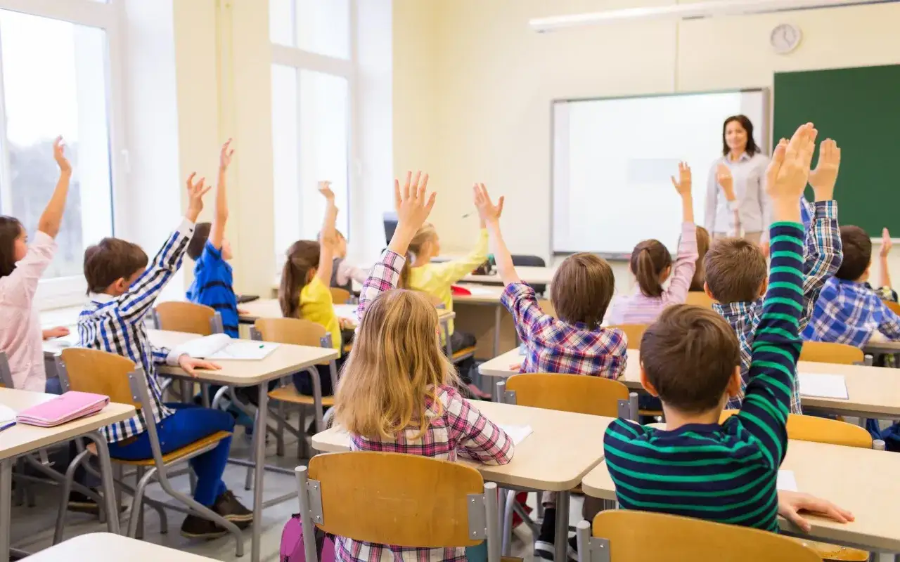 Uczniowie z podniesionymi rękami, czekający na odpowiedź nauczycielki. To obrazek z lekcji, która może być prowadzona zgodnie z najnowszym rozporządzeniem ministra edukacji narodowej.