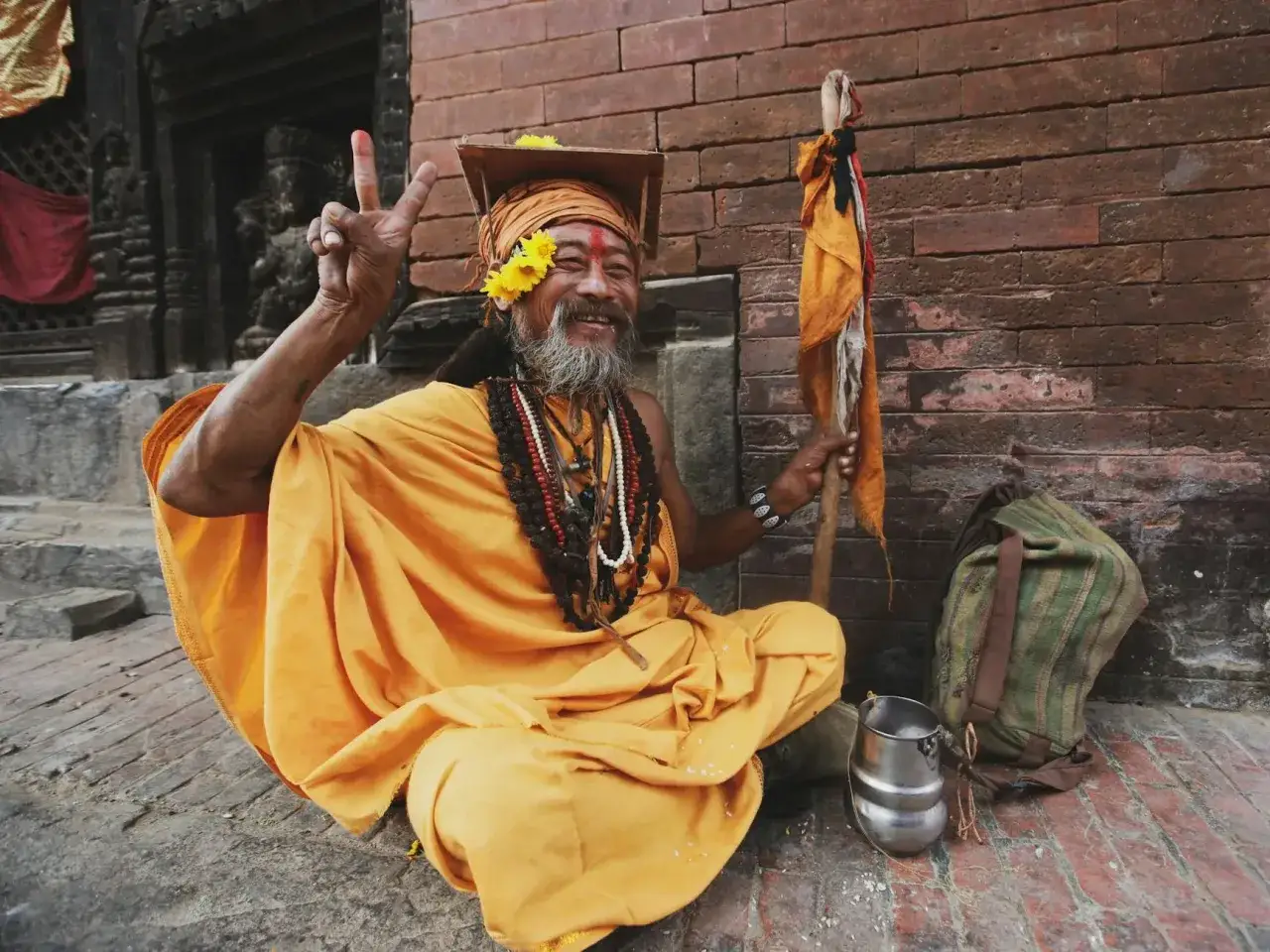 Uśmiechnięty sadhu w pomarańczowej szacie, z kwiatami we włosach, pokazuje znak pokoju. Jego postawa i skromne przedmioty wokół podkreślają, że asceza jako wartość jest jego drogą.
