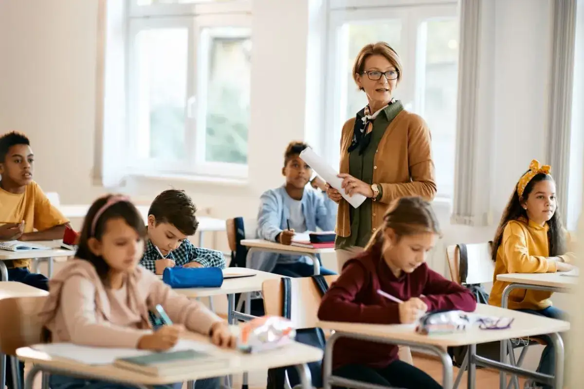 Nauczycielka z dokumentami prowadzi lekcję dla uczniów w 3 klasie liceum. Dzieci siedzą przy ławkach i piszą.