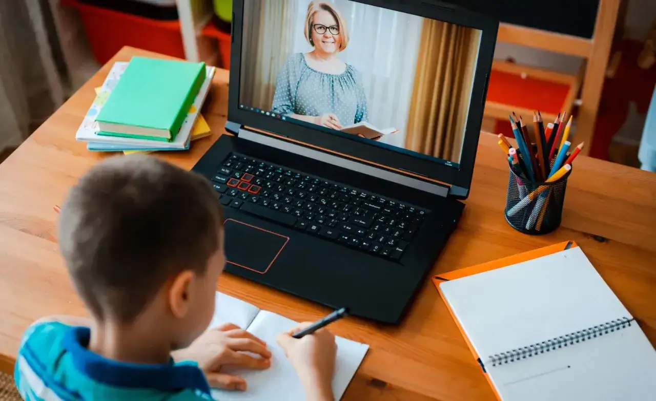 Chłopiec pisze w zeszycie podczas lekcji online. Na ekranie laptopa nauczycielka, co symbolizuje decyzję o nauce zdalnej.