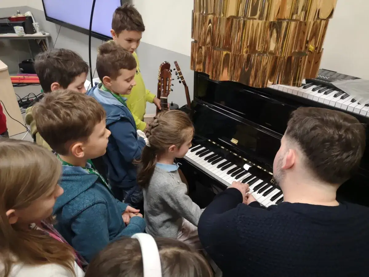 Dzieci słuchają lekcji muzyki, zastanawiając się, do której klasy jest muzyka. Nauczyciel gra na pianinie.